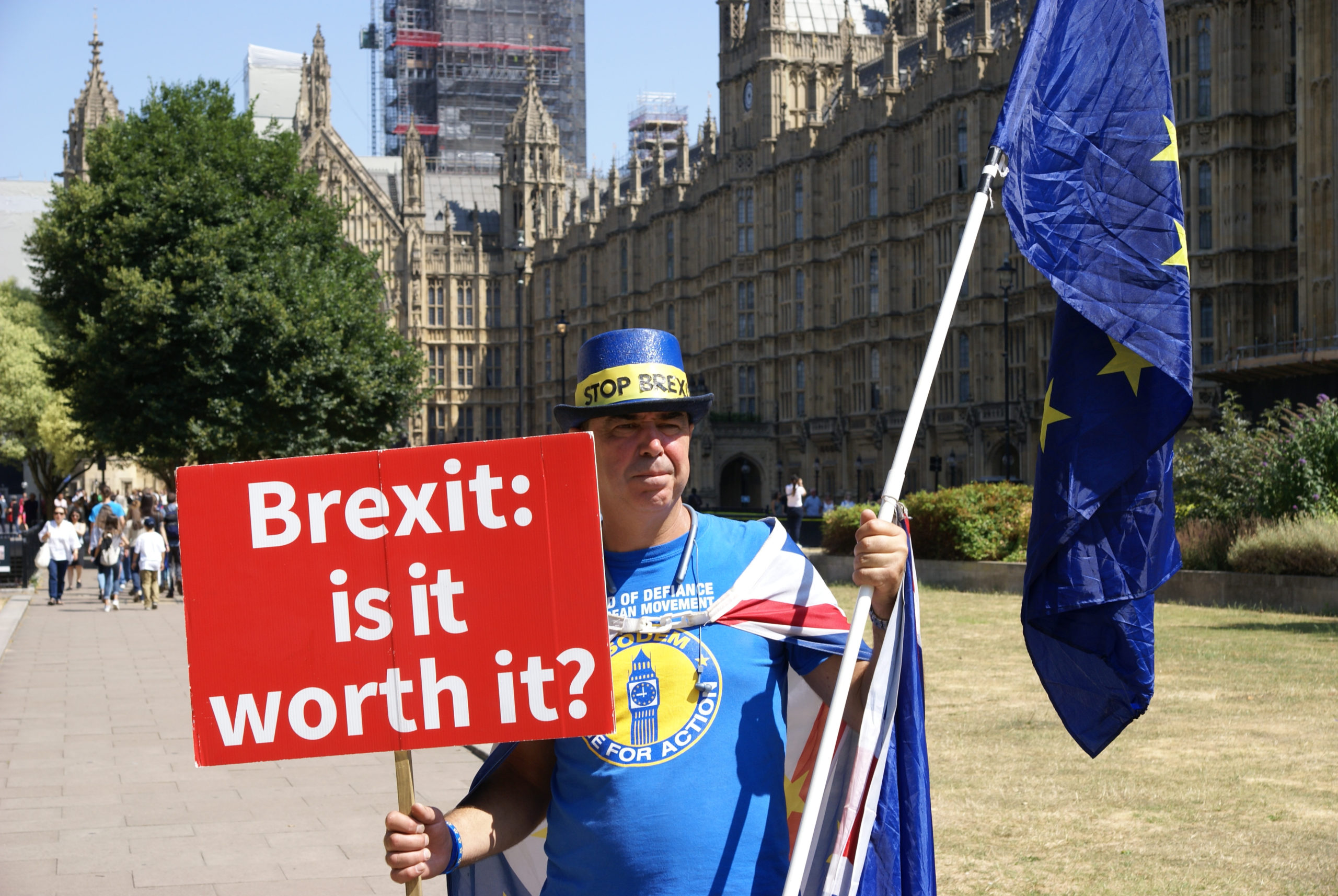 Mann kledd i blått med EU flagg står foran det britiske parlamentet med en plakat hvor det står "Brexit: is it worth it?"