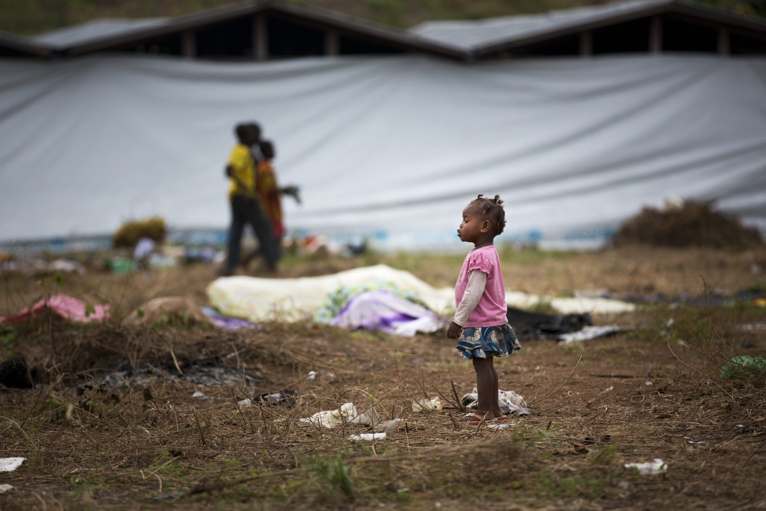 en liten jente står midt i en flyktningleir, telt i bakgrunnen
