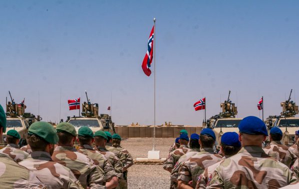 Soldater står oppstilt ute. Grus på bakken. Norsk flagg i midten. Militærkjøretøy står parkert. Flere norske flagg.