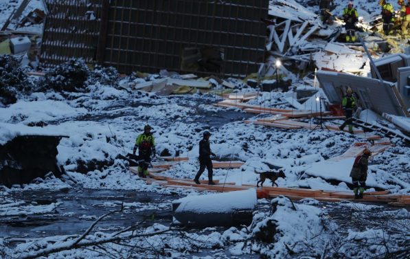 Rasområde i Gjerdrum. Dekket i snø og vann, samt bygningsdeler. Fire personer og en hund går på en bro laget av planker. I et av hjørnene kan man skimte personer fra redningsertater som lyser med lykter.