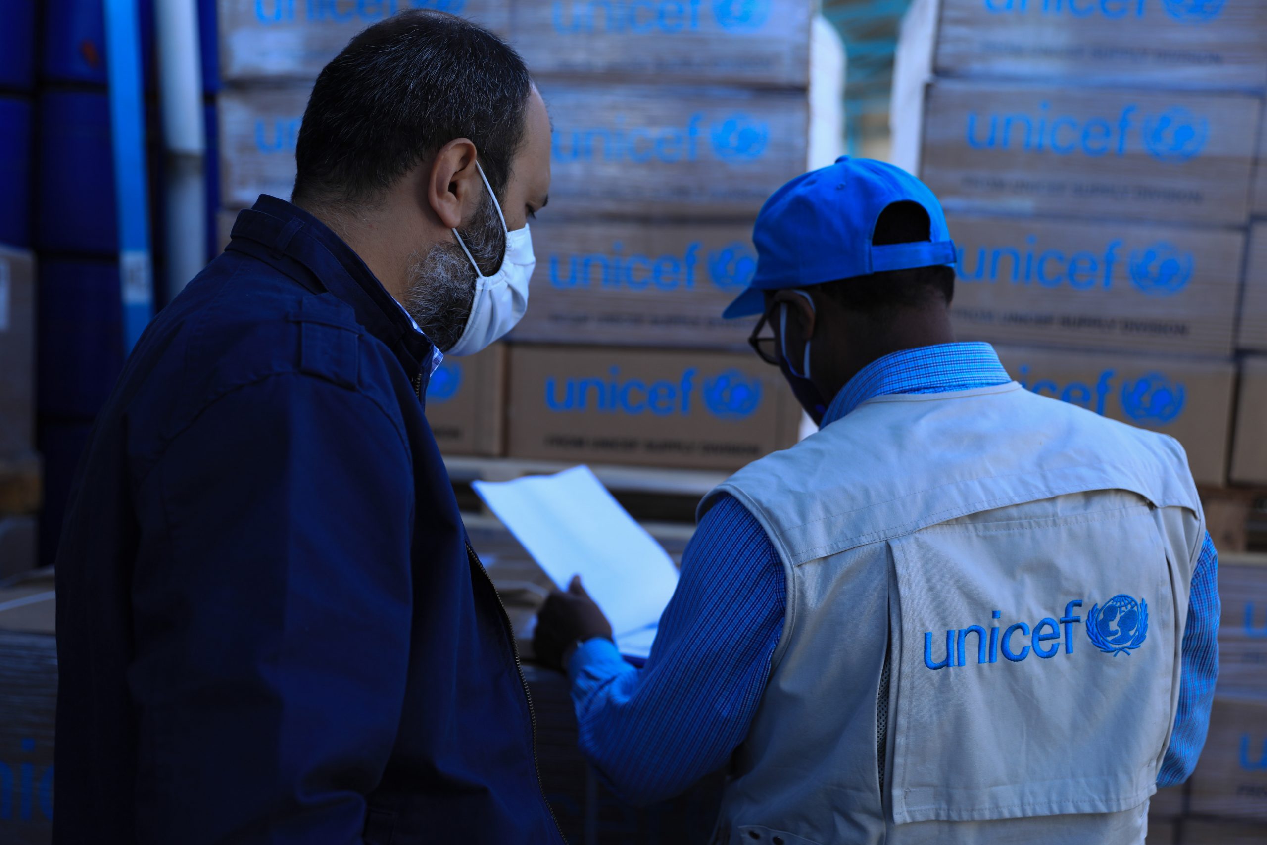 To personer ser på en liste, bak dem står det titalls pappesker hvor det står "UNICEF"