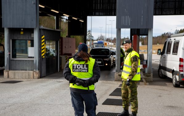 En person fra tollvesenet og en person fra heimevernet står og snakker sammen ved grenseovergangen på Svinesund. I bakgrunnen kan man se bilder i kø.