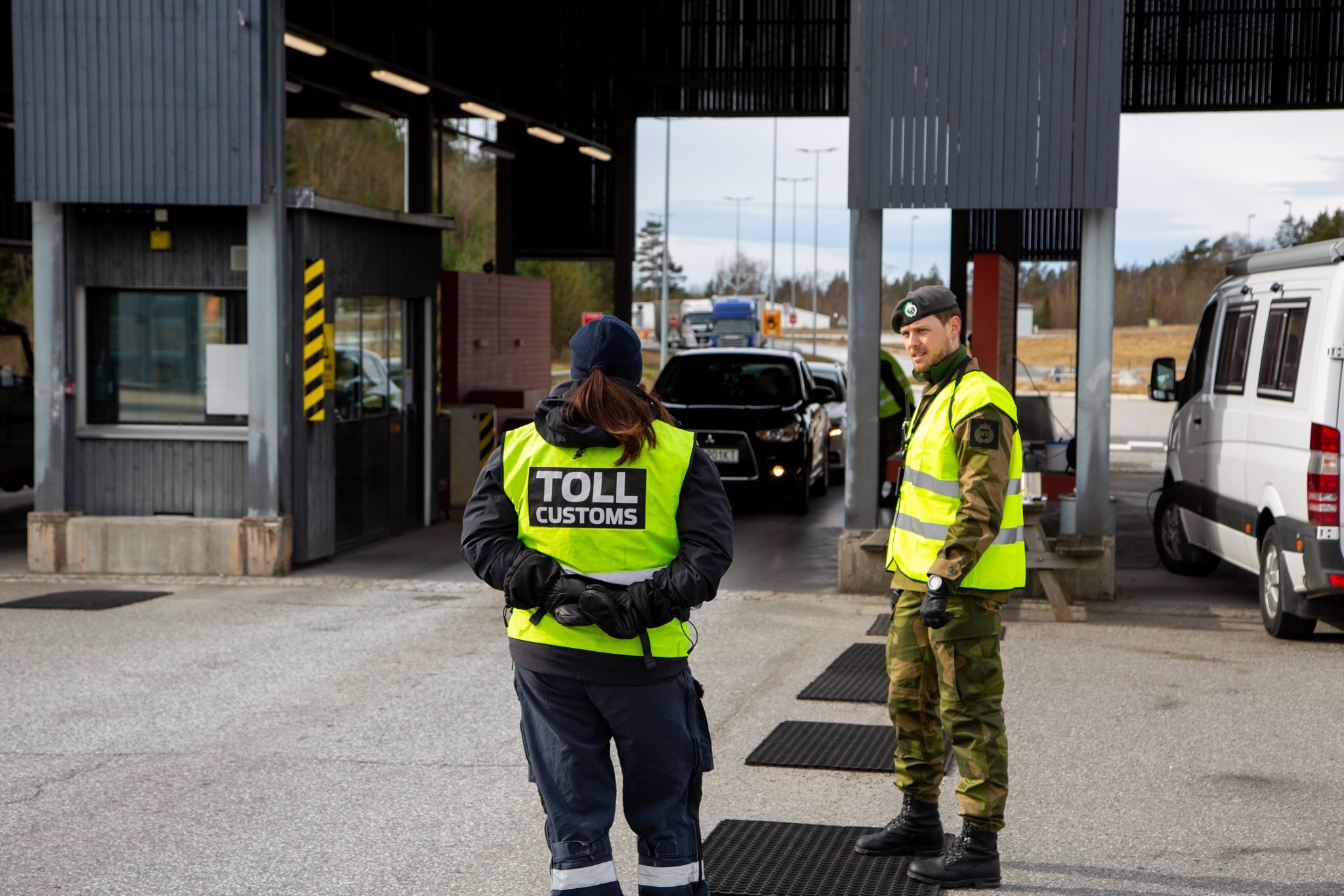 En person fra tollvesenet og en person fra heimevernet står og snakker sammen ved grenseovergangen på Svinesund. I bakgrunnen kan man se bilder i kø.