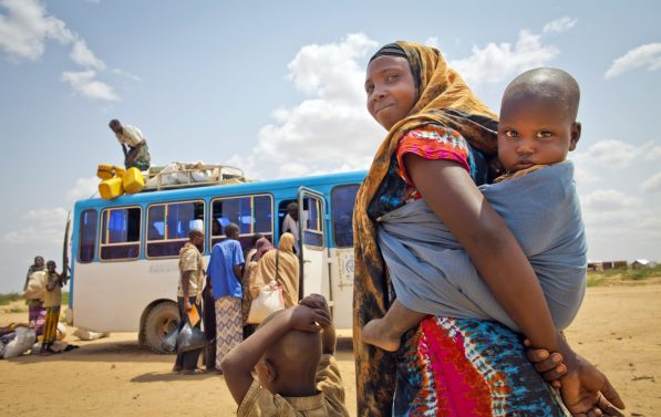 En kvinne med et barn på ryggen og et lite barn foran seg ser mot kamera. I bakgrunnen ser vi en buss som lastes.