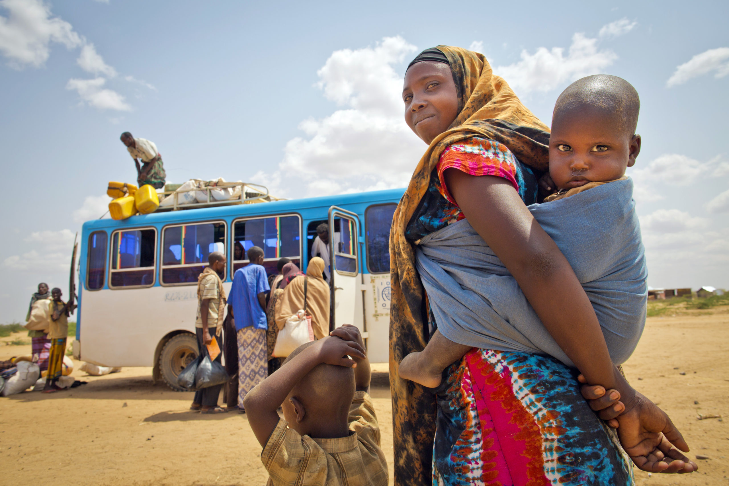 En kvinne med et barn på ryggen og et lite barn foran seg ser mot kamera. I bakgrunnen ser vi en buss som lastes.