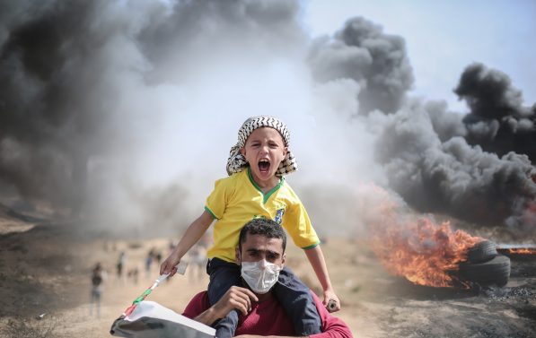 Et barn med palestinaskjerf på hodet, sitter på skuldrene til en mann. I bakgrunnen ses svart røyk.