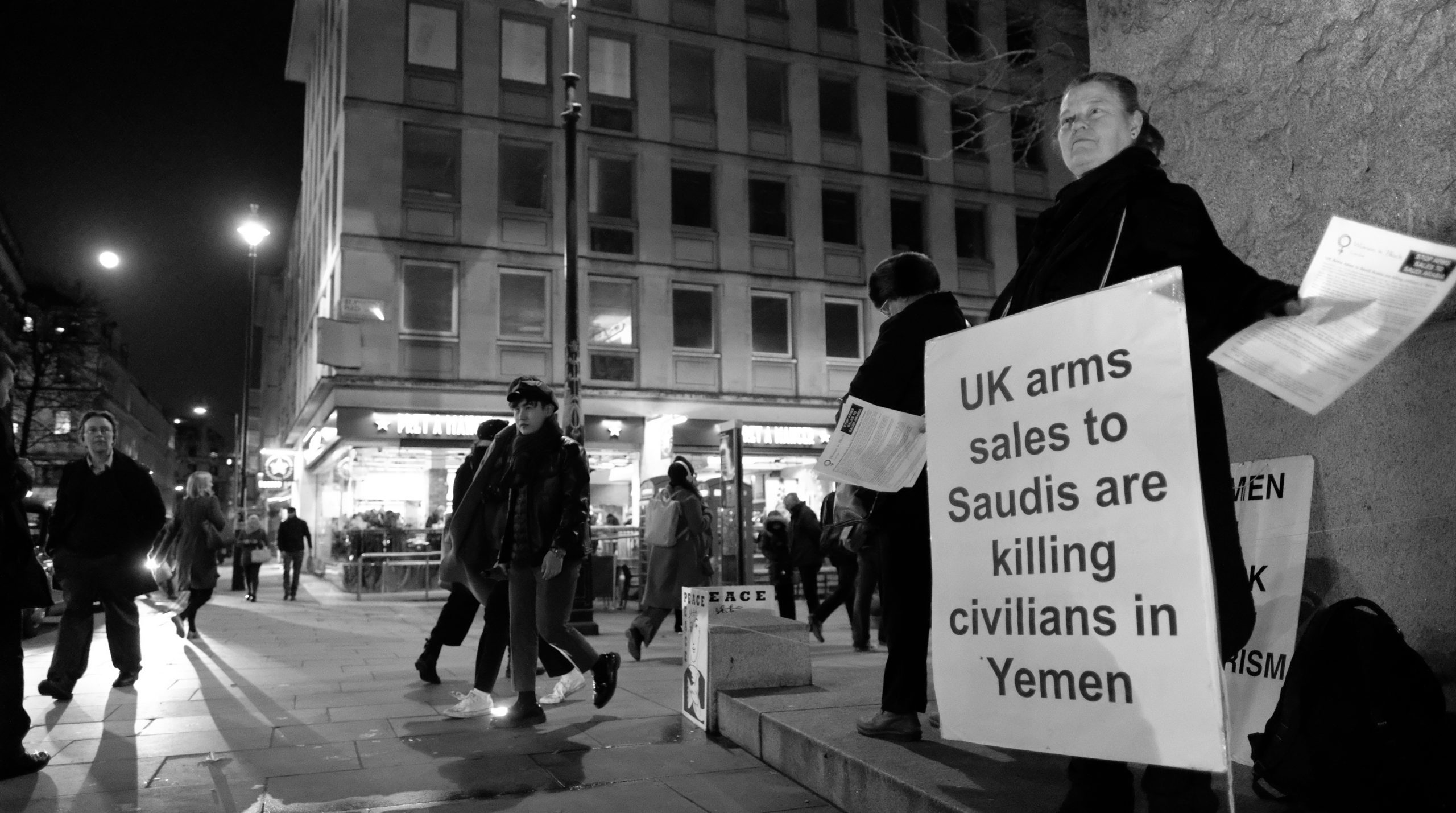 Svart-hvitt bilde av en kvinne som står på gaten med et skilt som informerer om hva britisk våpensalg fører til i Jemen. Hun deler ut flygeblader til forbipasserende.