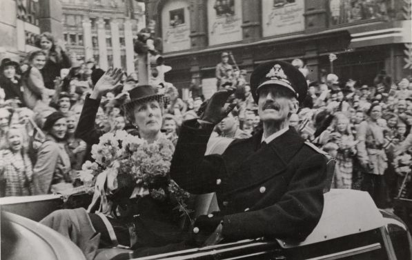 Kong Haakon og Dronningen sitter i en åpen bil og vinker til alle menneskene i gatene. Det er hundrevis av mennesker oppstilt i gatene med norske flagg og hender i været.