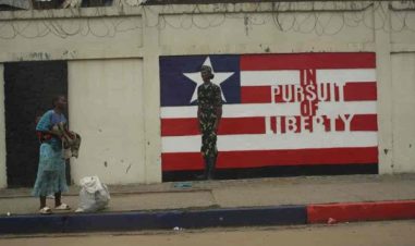 En dame står med sekk på gaten. Bak henne er det et bilde av flagget hvor det står "In Pursuit of Liberty"