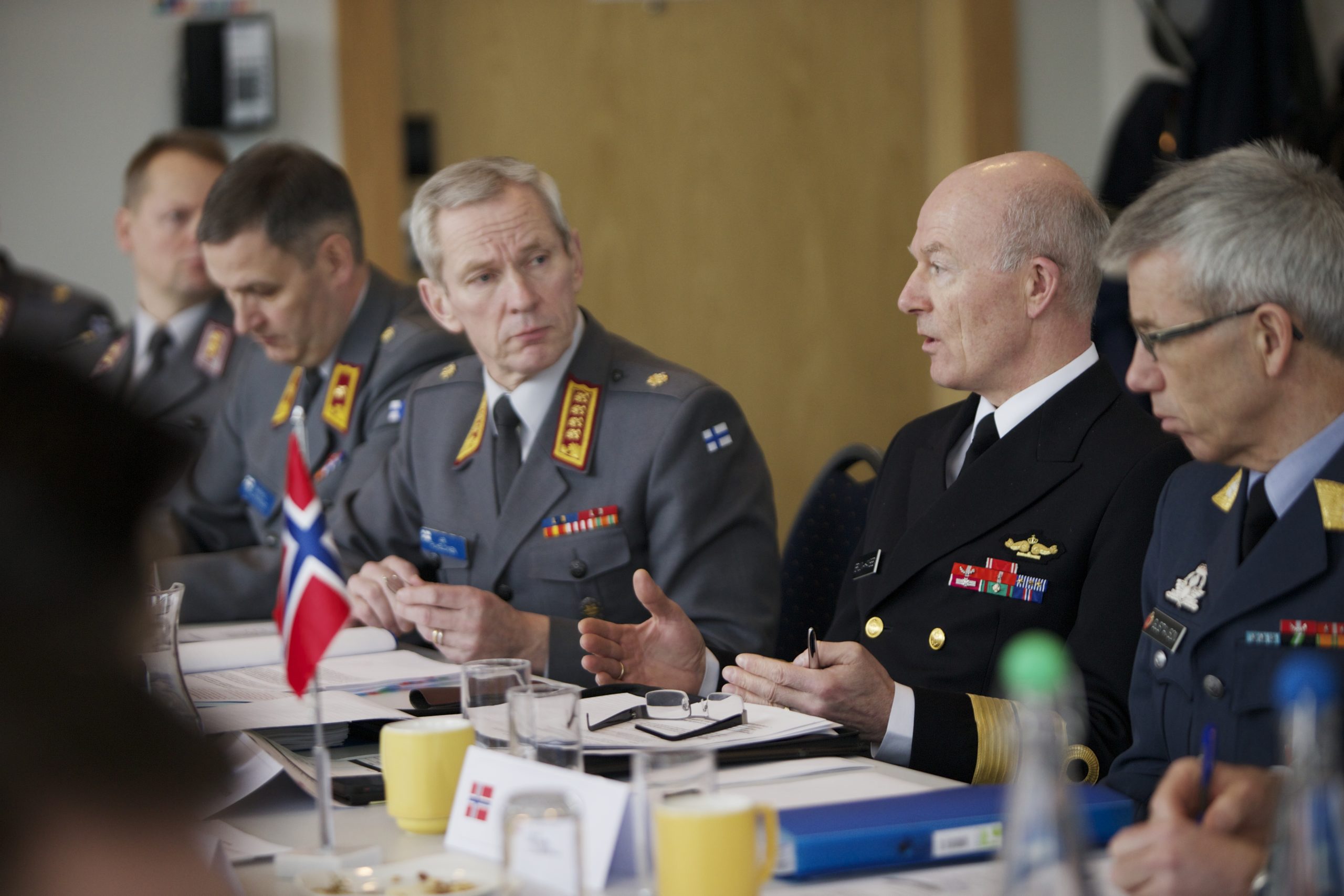 To menn kledd i uniform sitter ved et bord der blant annet det norske flagg står plassert.