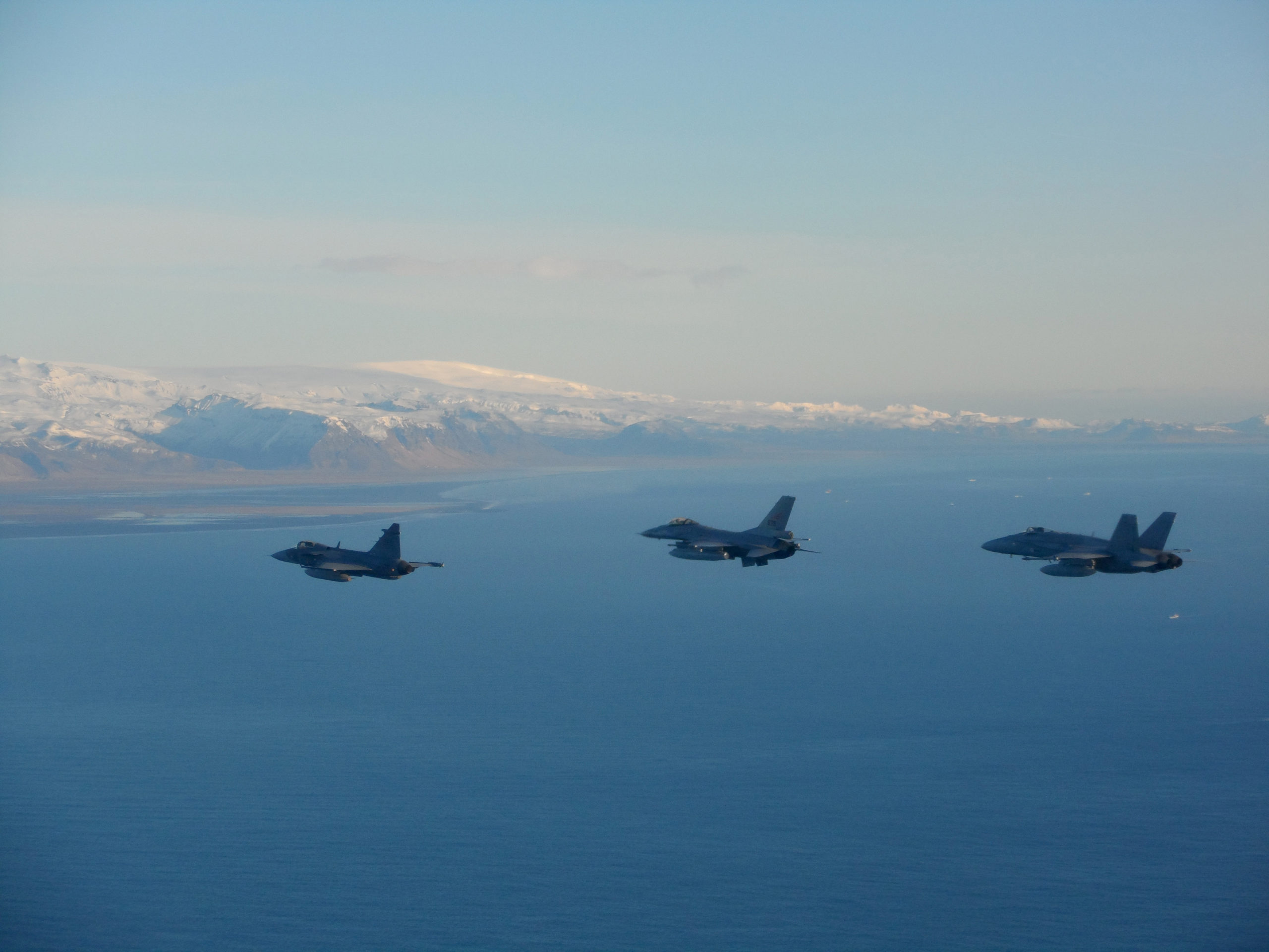 Tre jagerfly i formasjon over havet med snøkledde fjell i bakgrunnen.