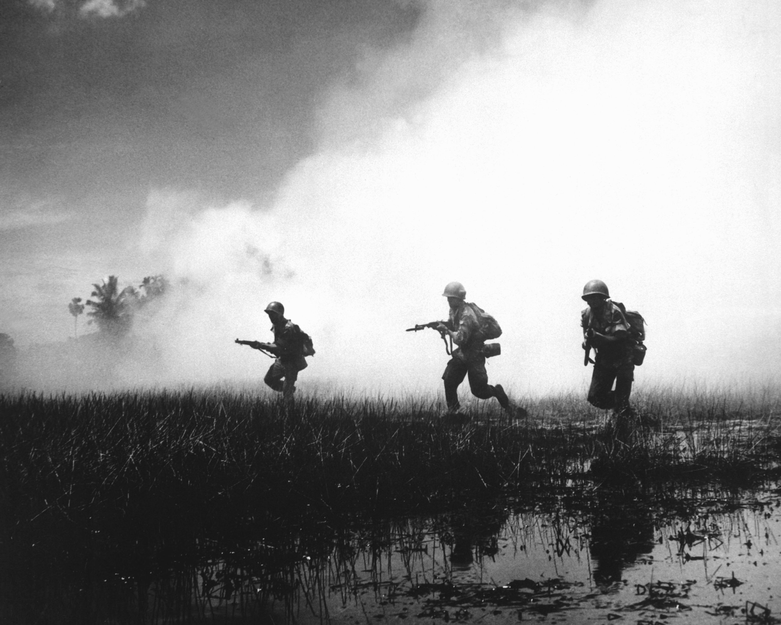 Tre soldater beveger seg bortover en sump med røyk i bakgrunnen.