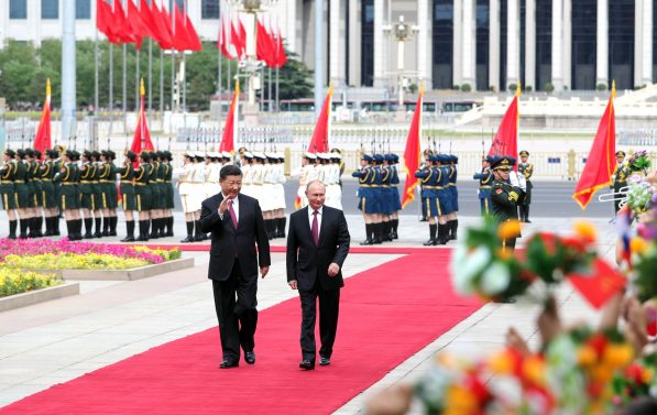 President Putin og President Jinping går nedover en rød løper og vinker til publikum. I bakgrunnen står militæret oppstilt.