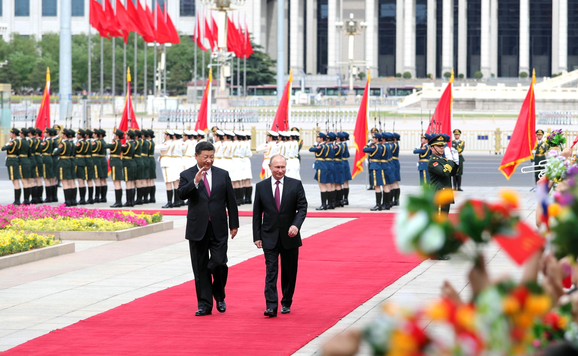 President Putin og President Jinping går nedover en rød løper og vinker til publikum. I bakgrunnen står militæret oppstilt.