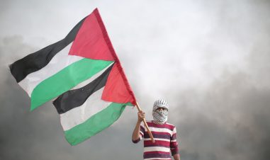 Mann med palestinaskjerf surret rundt hodet og ansikt holder to palestinaskjerf. I Bakgrunnen er grå røyk.