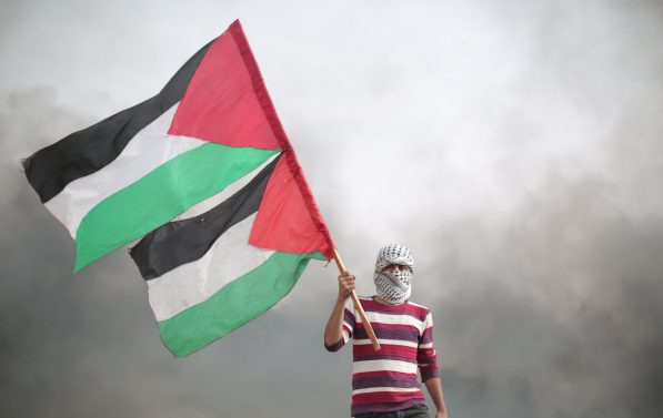 Mann med palestinaskjerf surret rundt hodet og ansikt holder to palestinaskjerf. I Bakgrunnen er grå røyk.