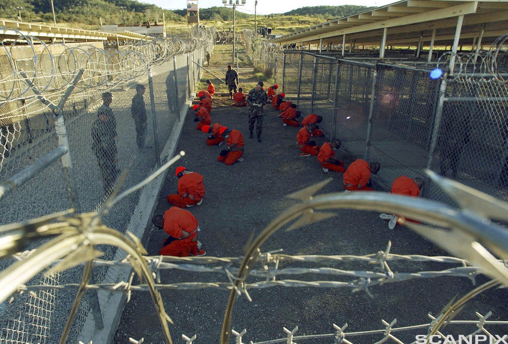 Fanger kledd i oransje uniformer sitter på asfalt med væpnede vakter rundt. Høye gjerder og piggtråd omringer dem.