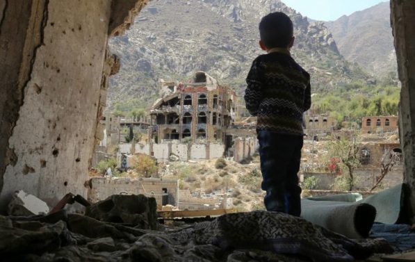 En liten gutt står i ruinene av en bygning og ser ut.