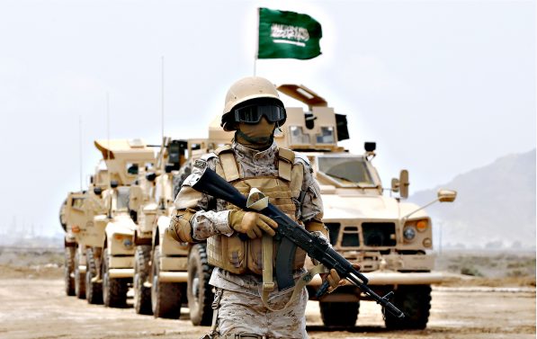 En soldat med våpen står foran en kolonne med bilder med det saudiske flagget på toppen