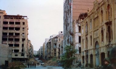 Ødelagte bygninger, soldater går langs veien i midten.