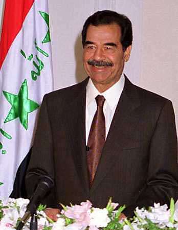 Saddam Hussein smiler ved et bord, i bakgrunnen er det irakiske flagget