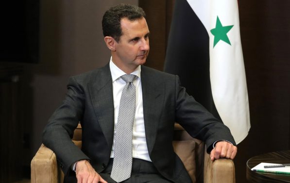 Bashar al Assad sitter i en stol. I bakgrunnen er det syriske flagget.