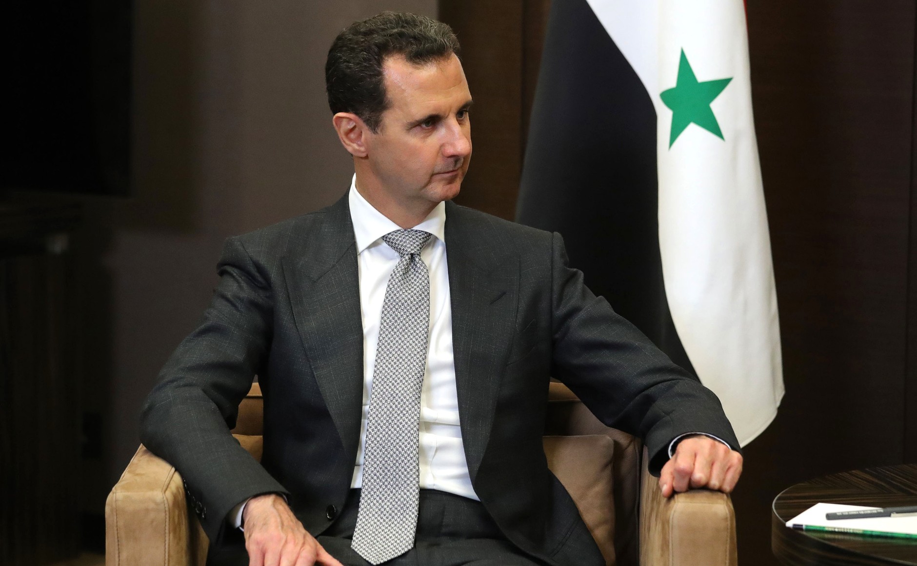 Bashar al Assad sitter i en stol. I bakgrunnen er det syriske flagget.