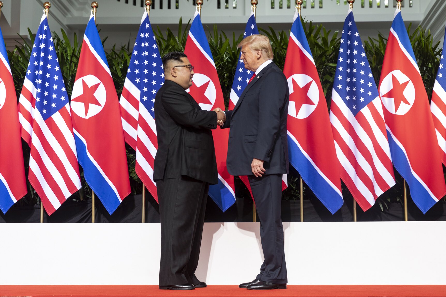 Trump og Kim Jung-un tar hverandre i hendene foran en rekke nord-koreanske og amerikanske flagg
