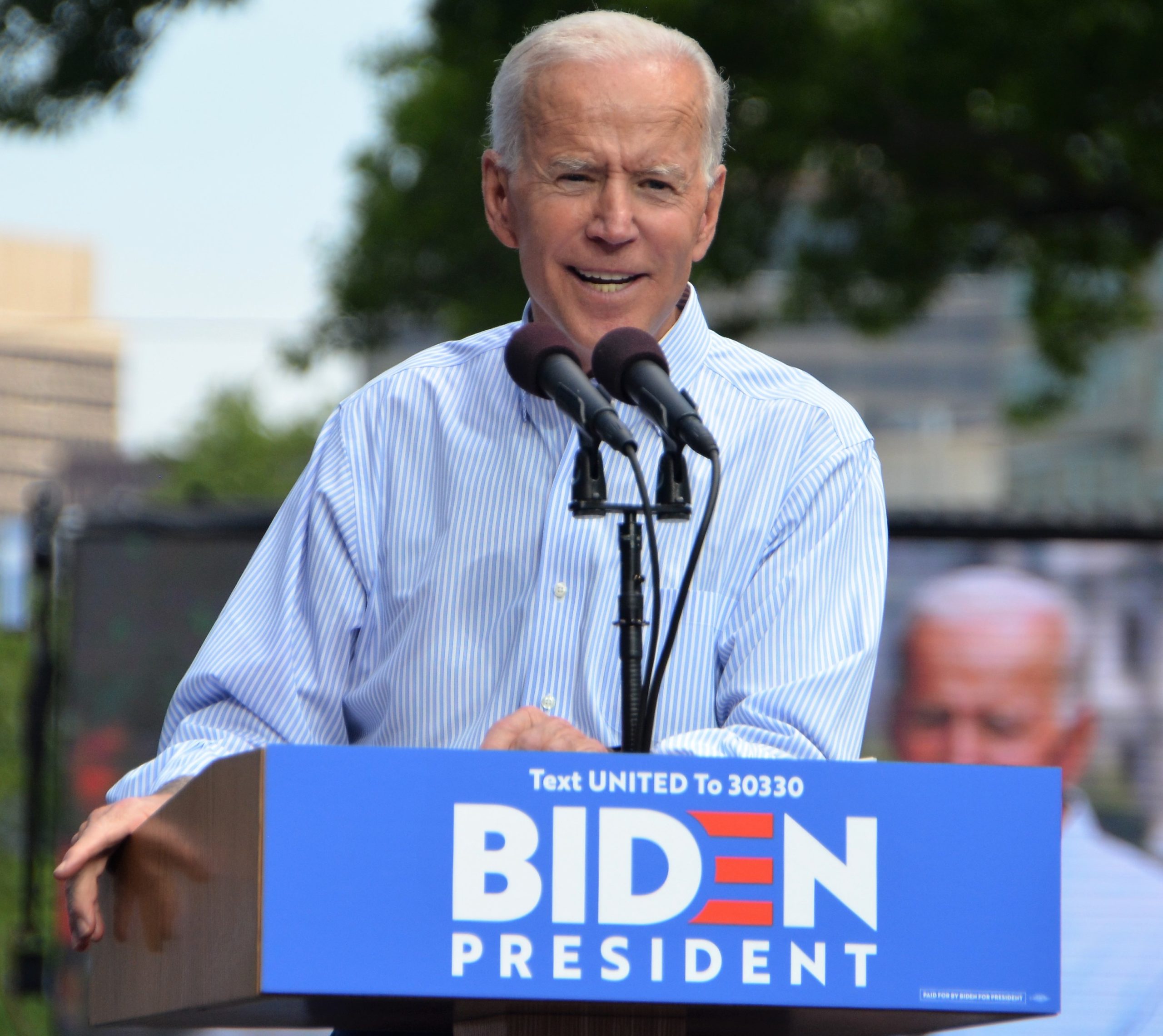 Joe Biden står ved en talerstol ute kledd i blå skjorte.