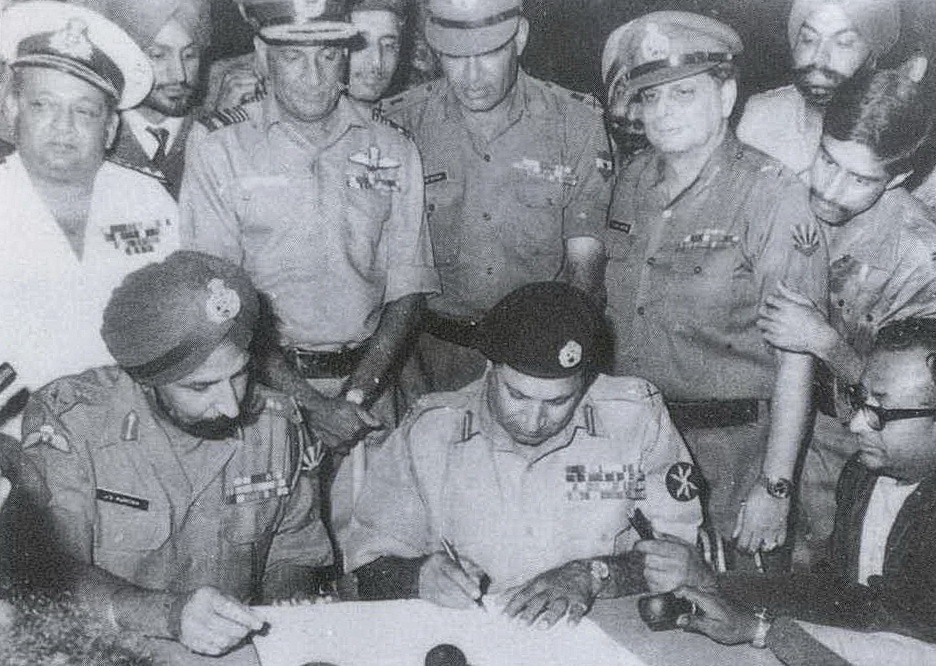 Tre personer sitter rundt et bord, mens 8 andre står rundt og ser på mens mannen på midten av bordet signerer et ark. 