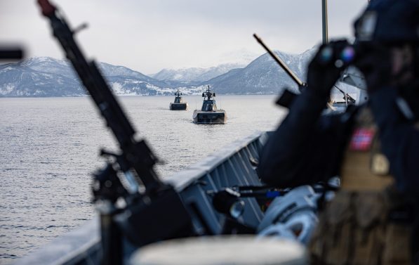 En soldat står på dekk av et skip og ser i gjennom en kikkert. Ved siden av hen står det våpen. I bakgrunnen kan man se to skip.