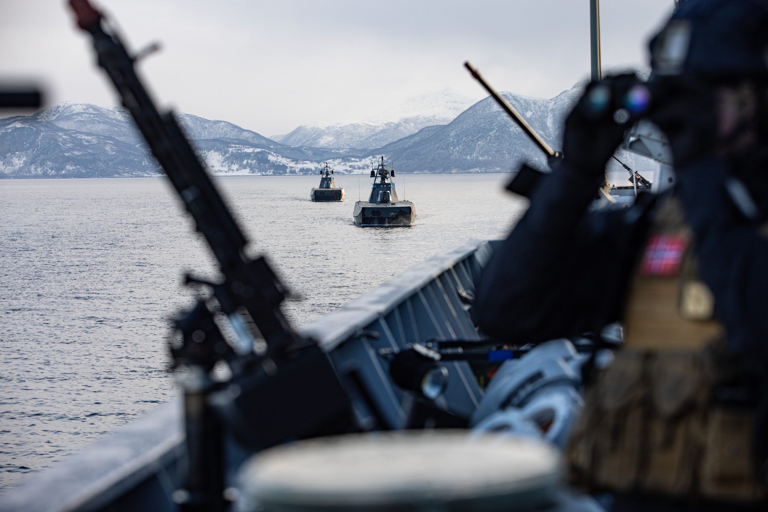 En soldat står på dekk av et skip og ser i gjennom en kikkert. Ved siden av hen står det våpen. I bakgrunnen kan man se to skip.