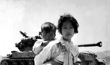 En foreldreløs koreansk jente bærer sin lillebror i et tørkle på ryggen. BAk henne står en militær tanks. Begge barna ser rett i kamera