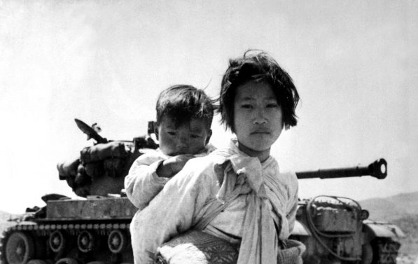 En foreldreløs koreansk jente bærer sin lillebror i et tørkle på ryggen. BAk henne står en militær tanks. Begge barna ser rett i kamera