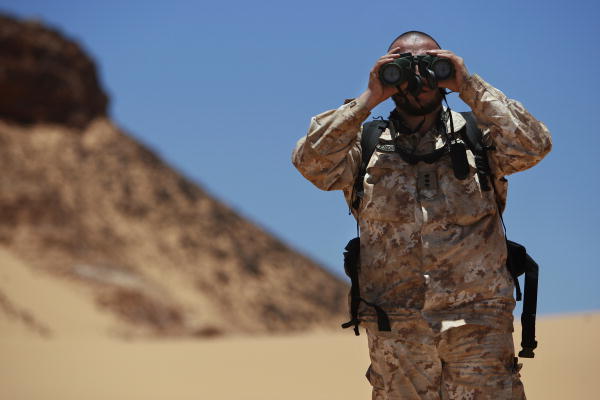 en mann står i en ørken med uniform på og ser i en kikkert