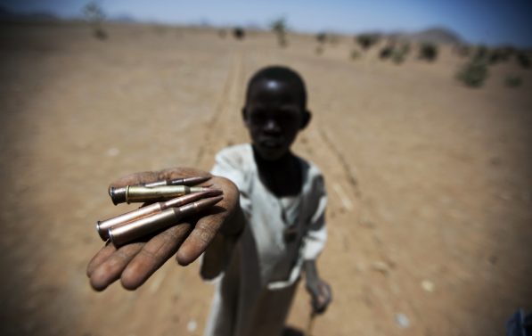 Et barn står ute i et tørt landskap og holder hånden mot kamera. I hånden har han våpen patroner