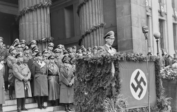 En mann taler fra en talerstol. På talerstolen henger nazistens hakekors. Bak ham stpr uniformerte menn
