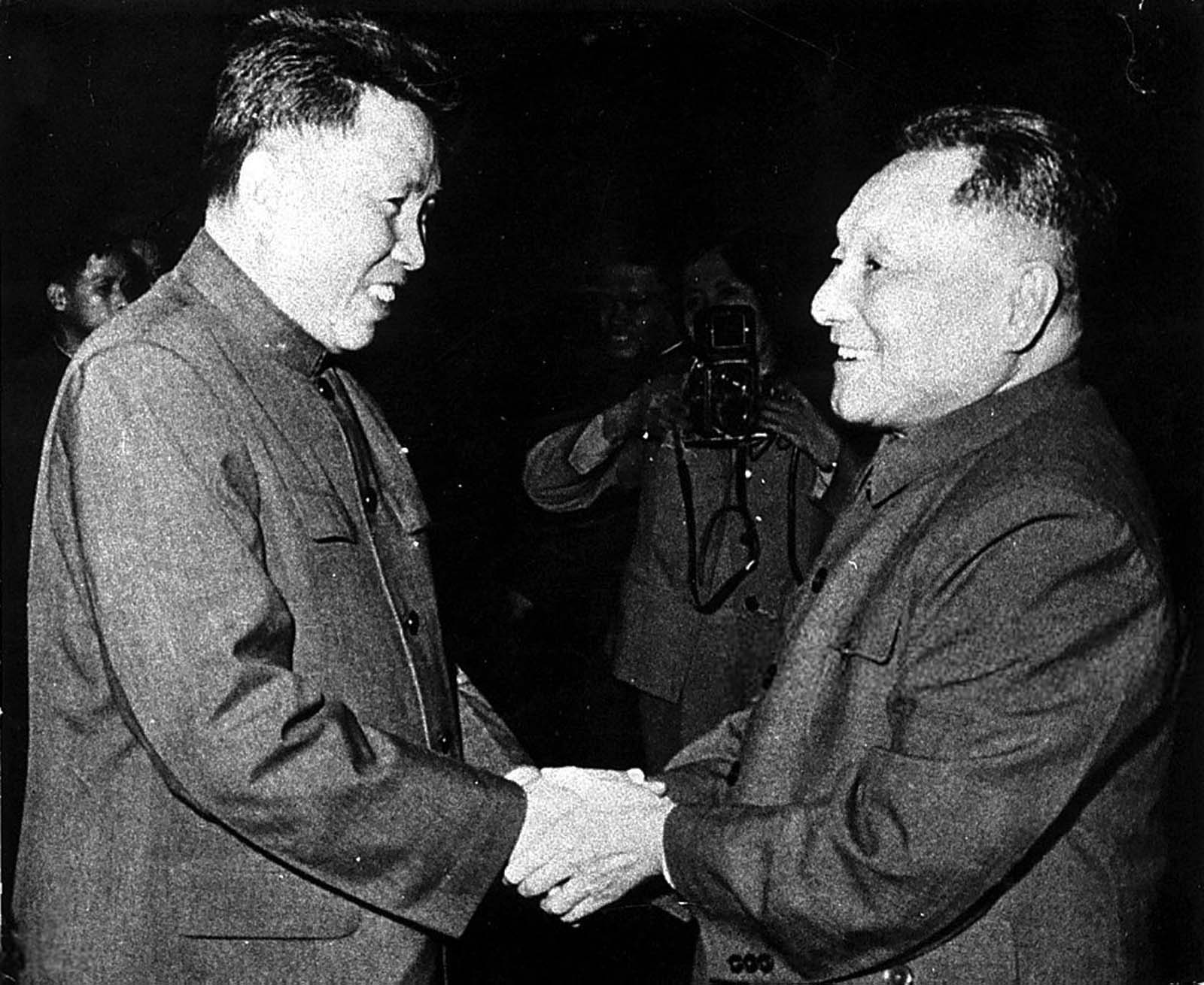 Pol Pot og Deng Xiaoping holder begge hender sammen, og smiler til hverandre. En fotograf tar bilde i bakgrunnen.