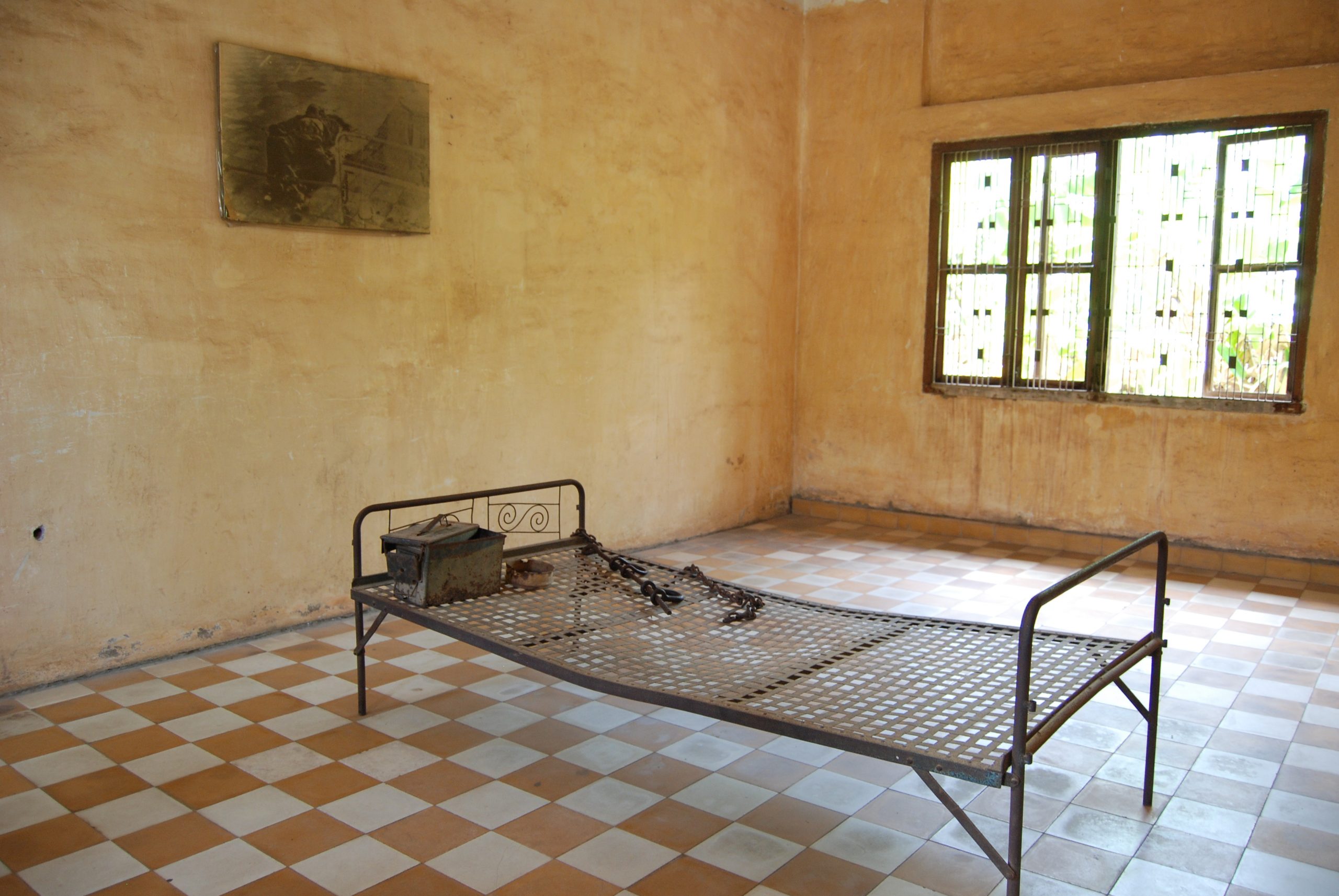 En metallseng med lenker står plassert midt i et gammelt klasserom med flislagt gulv i oransje og hvitt. En metallboks står plassert oppå sengen.