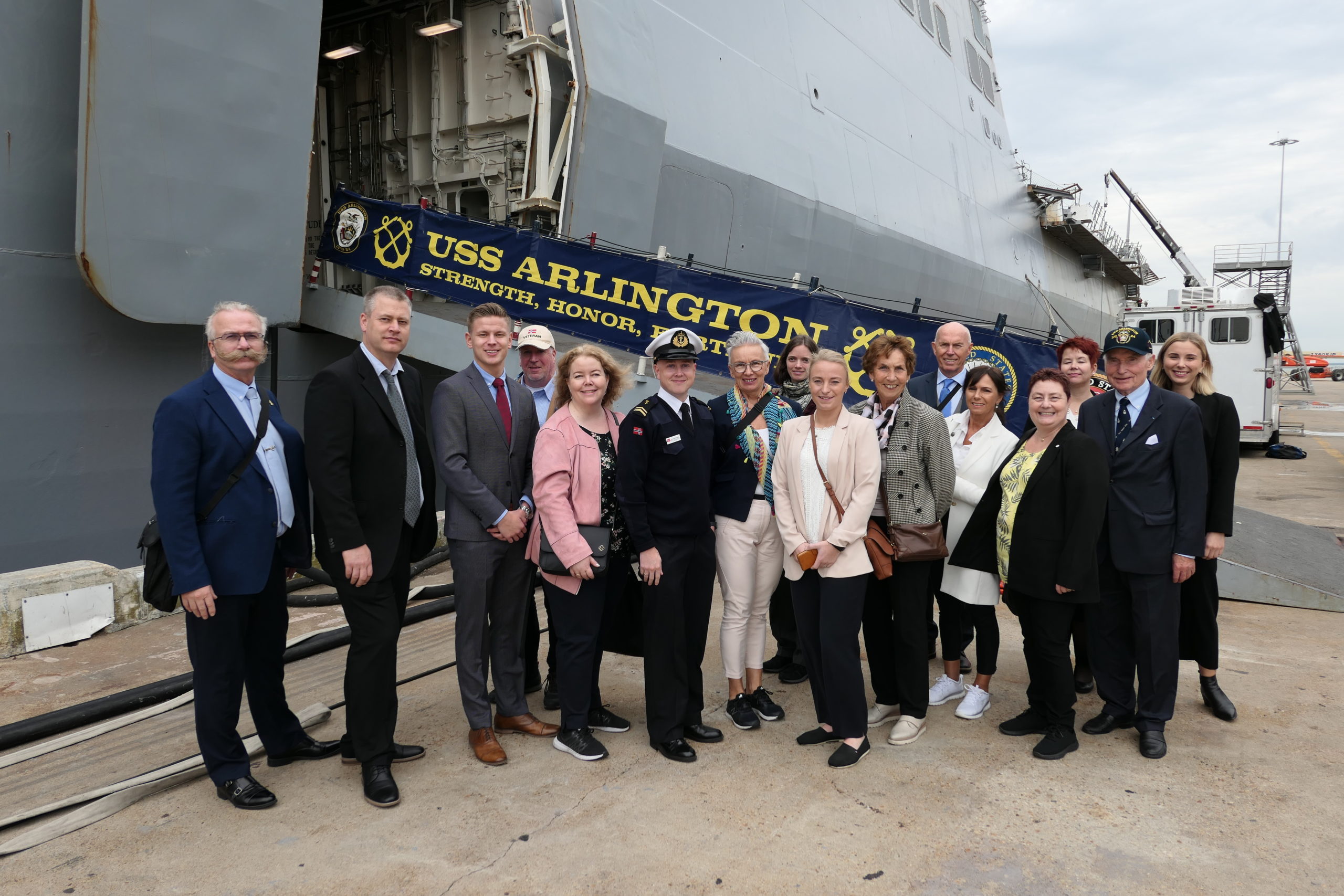 Gruppe poserer foran inngangen til et skip der man kan se USS Arlington skrevet på rekkverket