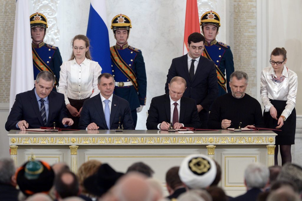 Tre personer sitter ved et bord, Putin i midten. Han og personen til høyre signerer et dokument. Bak dem står en kvinne og to soldater i uniform. Det er flagg på vaggen. Forran bordet sitter det masse folk på stoler.
