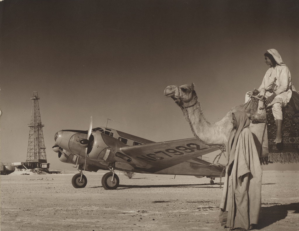 to menn, en på kamel, står foran et lite propellfly. I bakgrunnen ses en oljeinstallasjon