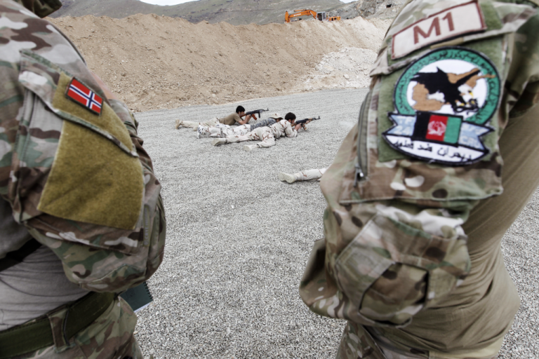 To soldater står i forgrunnen, på den ene kan du se et norsk flagg på skulderen. I bakgrunnen ser du to soldater som øver på å skyte.