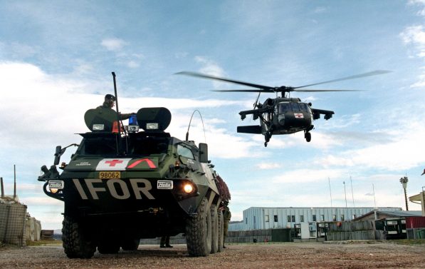 militært kjøretøy merket med IFOR bak står foran et helikopter som holder på å lande