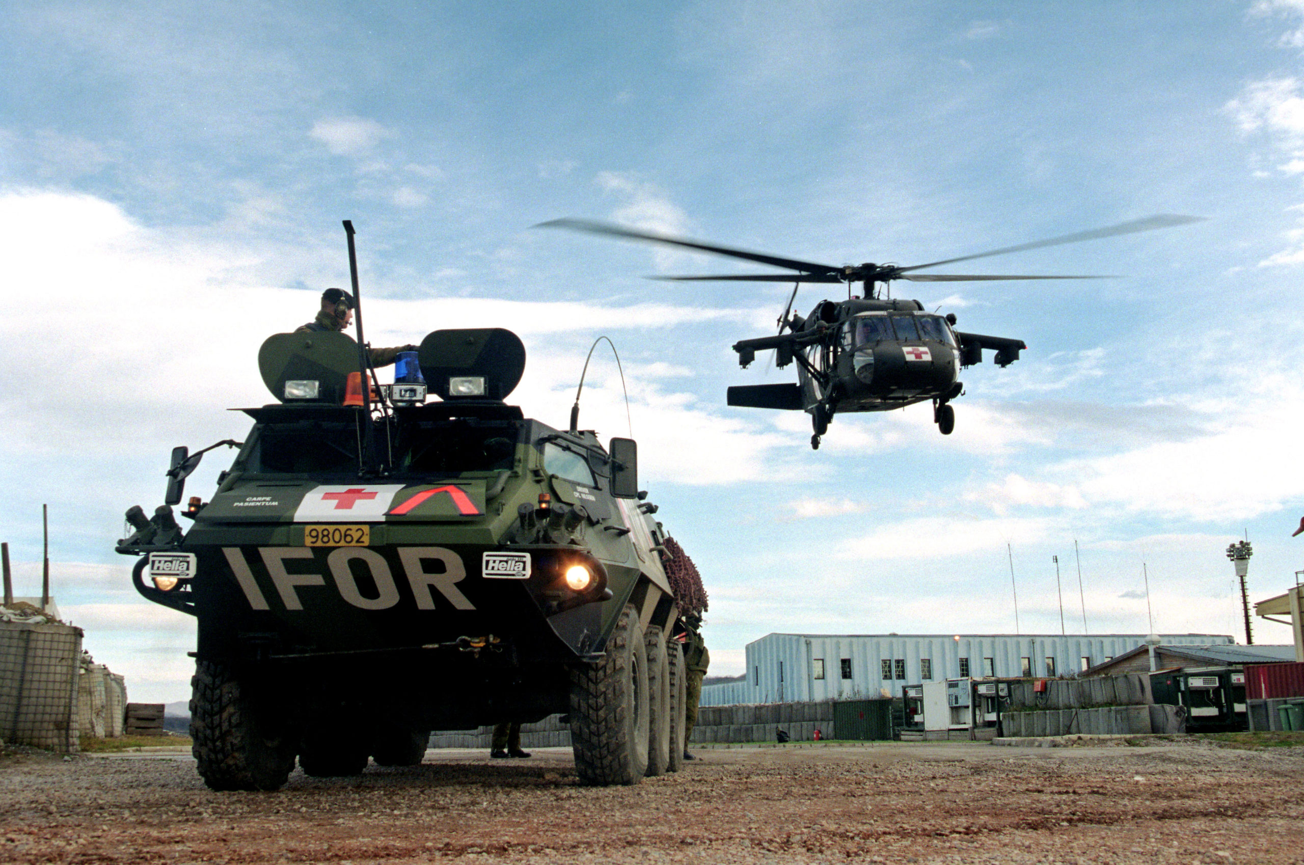 militært kjøretøy merket med IFOR bak står foran et helikopter som holder på å lande