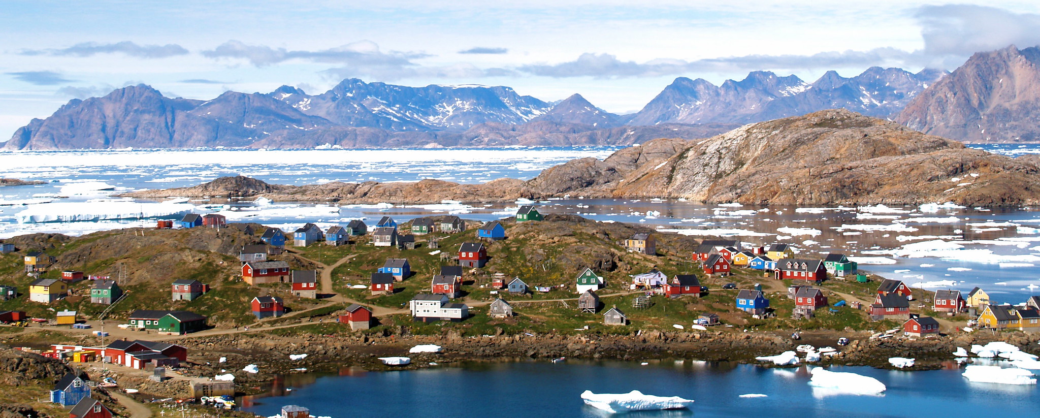 Hus ligger spredt på et landområde. I bakgrunnen ses høye fjell, og havet rundt er delvis dekket av is.