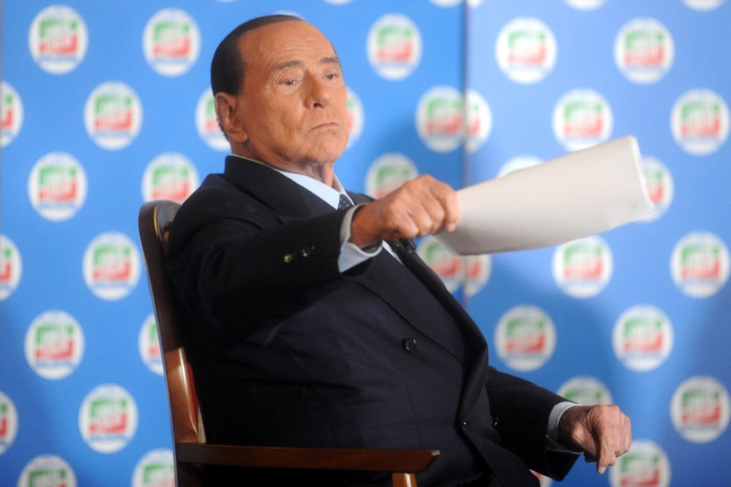 Berlusconi sitter i en stol og peker på noen med et ark.