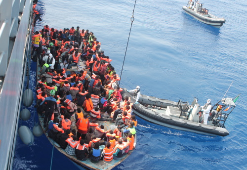 Båt overfylt med mennesker med redningsvester. Båten ligger inntil et større skip.