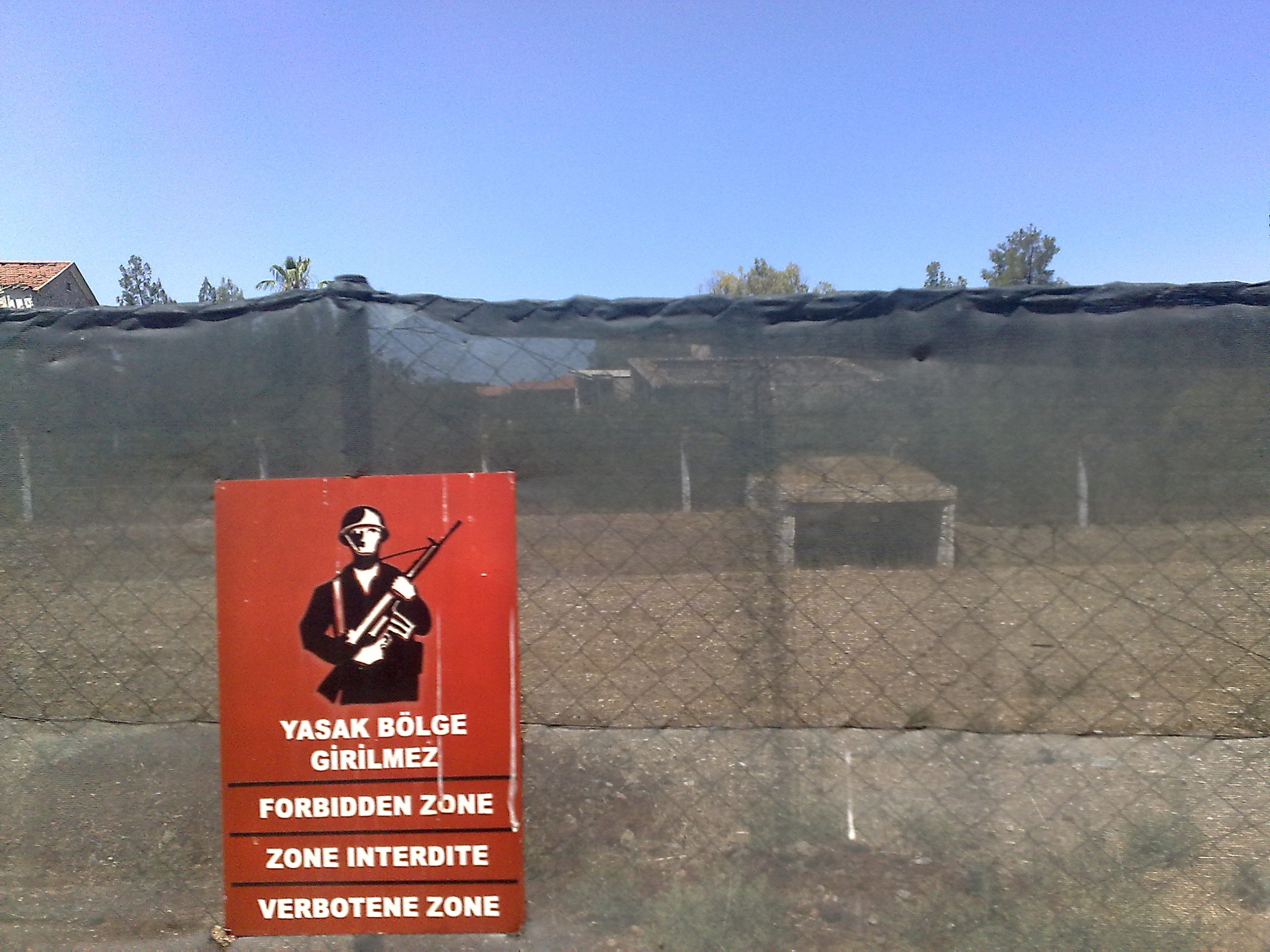 Et gjerde med en plakat i rødt der det er bilde av en soldat og det står "forbudt sone" på ulike språk.
