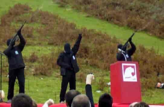 Tre maskerte personer kledd i svart poserer med våpen på et podium.